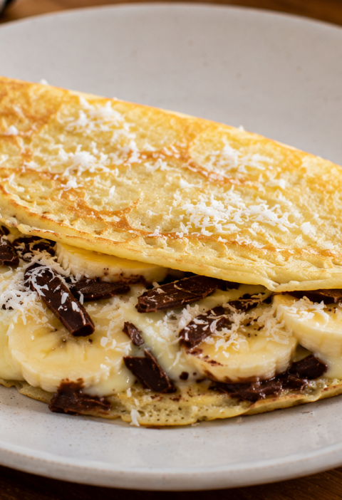 Crepioca recheada com banana, queijo derretido, coco e chocolate servida em prato caseiro